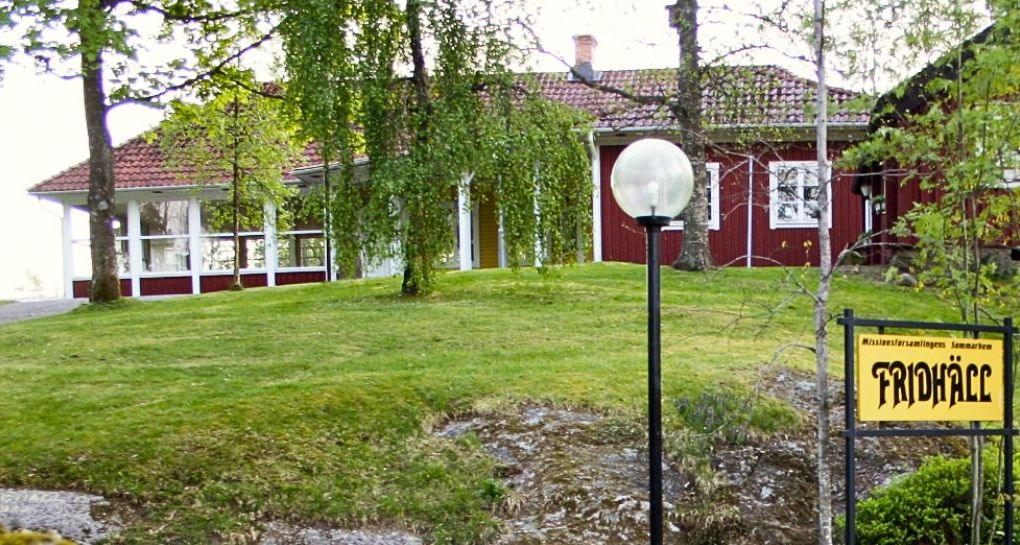 Sommarhemmet Fridhäll i rött i bakgrunden, med den gula skylten "Fridhäll" i förgrunden.