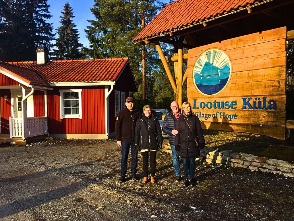 Fyra personer står framför ett rött hus till vänster och en stor skylt till höger där det står "hoppets by" på estniska och engelska.